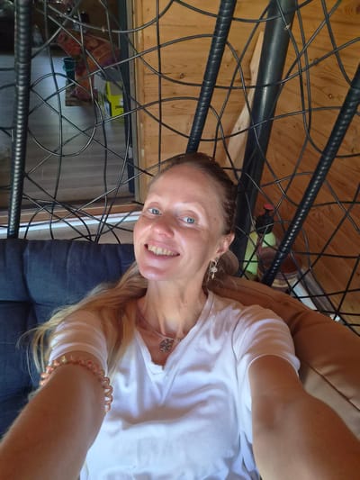 An adult female is depicted in a selfie, seated in a dark-framed, woven-style swing chair with dark grey and tan cushions, in an indoor setting located in Motyakovo, Russia. She has fair skin, blue eyes, and light blonde hair pulled back. Her expression is a broad smile, revealing her teeth. She wears a white V-neck t-shirt, a silver-colored necklace with an ornate circular pendant, and multiple earrings on her right ear, including a small hoop and dangle charms resembling a tree of life and a feather. On her left wrist, she wears a light-colored spiral hair tie or bracelet. The background behind her features light-toned horizontal wooden paneling walls and grey-toned tiled flooring. Visible through the chair's frame are various objects: a dark green bucket, a patterned fabric in red and purple tones, a yellow box with "JHAC" visible, a light green plastic bottle with a red pump top, and a dark brown container. A light wooden board is also partially visible in the upper right background.
