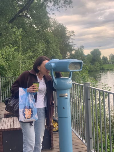 A young woman is using a public telescope overlooking a body of water. She is wearing a white t-shirt, blue jeans, and a brown jacket. She holds a cup in her right hand and a bag with a cartoon hedgehog graphic in her left hand. The telescope is light blue and mounted on a stand with a yellow wheel. The scene appears to be a park or recreational area with lush green trees and a wooden walkway. The sky is cloudy with hints of sunlight breaking through, suggesting it could be late afternoon or early evening. The location is Kolpino, Russia. There is no visible text in the image.