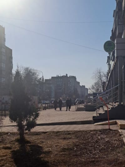The image displays an urban street scene in the city of Меткомелино, Russia, under clear, bright daylight with strong sun glare emanating from the upper left quadrant. The foreground features a patch of dry, sparse brown grass on the left with a small, dark conifer tree casting a pronounced shadow towards the viewer. To the right, a multi-story building exhibits a concrete staircase with silver metal railings and a single orange vertical support element. Affixed to this building's facade is a circular green sign bearing a dark, stylized emblem resembling a figure in a hat, positioned above a string of small, decorative lights.
A wide, paved pedestrian walkway, constructed from light-colored pavers, extends from the midground into the distance. Three individuals, silhouetted against the bright background, walk away from the viewer along this path. They appear to be adults dressed in dark clothing. Further down the path, additional indistinct figures are visible. Beyond the pedestrian area, a street is visible with parked cars. The background is dominated by a row of multi-story buildings on both sides of the street, receding into the horizon, characteristic of urban residential or commercial architecture. Bare deciduous trees and streetlights are interspersed throughout the midground and background. The sky is light blue and clear, traversed by two thin utility lines near the top of the frame.