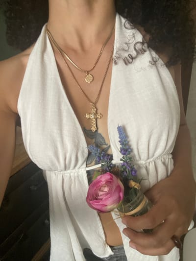 The media file shows a close-up of a person's upper body, focused on their chest and shoulder area. The person has dark, curly hair that falls over their left shoulder. They are wearing a white, ruched top that exposes their décolletage.

Adorning the person's chest are several pieces of jewelry. A delicate gold chain with a circular pendant rests on their collarbone, and another gold chain with a cross pendant is layered beneath it. A tattoo is visible on the person's chest, peeking out from beneath the top.

To the right of the cross pendant, a small bouquet of flowers is attached to the top. The bouquet consists of a large pink rose, some purple sprigs that appear to be lavender, and other green foliage. A hand, adorned with a ring on one finger, is resting over the bouquet and the side of the person's chest. The hand is holding a small glass jar, which is wrapped with twine.

The setting appears to be indoors, with a dark, wooden surface visible in the background. The lighting suggests it could be daytime, but no specific weather or time of day cues are strongly indicated. There is no visible text in the image. The overall mood conveyed is one of calm and perhaps a touch of bohemian or spiritual aesthetic due to the flowers and jewelry.