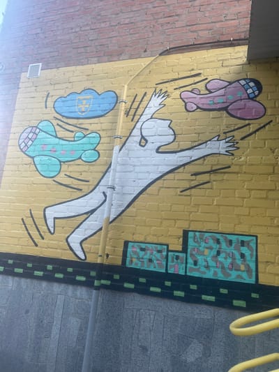 The image displays a mural painted on a yellow brick wall. The mural features a stylized white humanoid figure with arms outstretched, appearing to be in mid-air. Two cartoonish airplanes are depicted flying around the figure: one light blue with pink polka dots to the left, and one pink with light blue polka dots to the right. Above the figure, a blue cloud with a yellow emblem is visible. The bottom of the mural depicts a dark band with green squares, resembling a road or ground. The wall is topped by a section of red brick. A metal ventilation grate is present on the upper left portion of the yellow wall. A yellow pole, possibly a lamppost or part of a structure, runs vertically through the mural, partially obscuring the white figure. The ground below the mural is made of gray stone slabs. This mural is located in the city of Horodok, Ukraine.