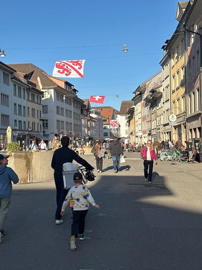 The image depicts a bustling street scene in Winterthur, Switzerland, under a clear, bright blue sky. The street is lined on both sides by multi-story buildings showcasing traditional European architecture, with various windows and facades. Long shadows stretch across the asphalt pavement, indicating a sunny day, likely during late morning or early afternoon.
Numerous people are visible on the street, walking and congregating. In the foreground on the right side, a person in a dark jacket stands beside a child. The child wears a light-colored long-sleeved shirt patterned with yellow Minions characters, dark pants, and a red bib on their chest displaying the numbers "26 78," suggesting participation in an organized event or race. Another individual in a blue hoodie and khaki pants is partially visible in the bottom right corner, looking upwards. Further down the street on the right, a woman in a bright pink jacket and dark trousers walks towards the viewer.
On the left side of the street, several bicycles are parked against a building, and more people are gathered, some appearing to be seated at outdoor establishments. Above the buildings, two flags are prominently displayed against the clear sky. One is the distinctive square red Swiss flag with a white cross. The other is a rectangular white flag featuring a red diagonal stripe and three red lions or griffins, resembling the flag of the Canton of Thurgau.
Visible text includes the numbers "26 78" on the child's bib. On a shop sign situated on one of the buildings on the left, the word "APPENZELLER" is clearly legible, with "Löwen" also faintly discernible, possibly indicating a restaurant or brewery. The overall atmosphere is lively and typical of an active town center.