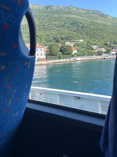The image is taken from inside a vehicle, likely a bus or ferry, looking out through a window. In the foreground, the blue patterned seat of the vehicle is visible on the left and a dark blue fabric is on the right. The middle ground features a body of water, which appears to be a bay or a strait, with gentle ripples on its surface. Along the far shore, there is a waterfront promenade with a stone wall. Behind the promenade, a town is visible, characterized by buildings with red-tiled roofs and white facades. Some of these buildings appear to be multi-story residences. Beyond the town, a densely forested green mountain rises steeply, with its peak reaching into a clear blue sky. The overall impression is a scenic view of a coastal town nestled against a mountainous backdrop, likely in a sunny, warm climate. The specific location in Đurići, Montenegro, suggests a view of the Bay of Kotor, a region known for its dramatic scenery where mountains meet the sea.