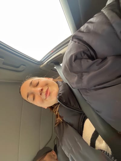 The image displays the interior of a vehicle, featuring one primary human subject and a partial view of another. The main subject, a female with brown hair, occupies the central-right portion of the frame. Her eyes are closed, and her mouth is slightly upturned in what appears to be a serene expression. She is wearing a dark, possibly black, puffer jacket over a cream-colored garment with black horizontal stripes. A dark seatbelt is fastened across her chest. Above her, an open sunroof reveals a bright, overcast sky. A subtle red line is discernible along the upper edge of the sunroof mechanism. In the lower-left corner, the top of another individual's head, partially obscured and wearing a dark cap, is visible. The vehicle's interior surfaces are primarily grey or beige. The image does not contain visual information to independently confirm the specific city location as Hoenheim, France.