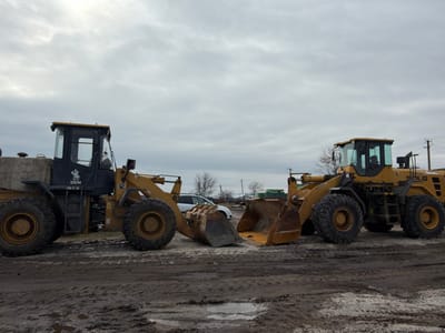 In Bessergenovka, Russia, an outdoor industrial or storage yard is depicted under an overcast, grey sky. The scene is dominated by two large yellow wheel loaders parked on muddy, uneven ground marked with tire tracks and small puddles.

On the left, a SEM brand wheel loader with a black cab and yellow body is positioned. It has large, dirty tires and a prominent front bucket. A sticker featuring a cartoon character is affixed to the cab door. Directly opposite, to the right, is another yellow wheel loader, identified by "SOLG" (likely SDLG) on its side. This loader also features large, dirty tires and a front bucket. The two loaders are positioned facing each other, with their front buckets nearly touching at ground level, suggesting they are parked in close proximity.

In the mid-ground, a white passenger vehicle is partially visible behind the left loader's bucket. In the background, particularly behind the right loader, utility poles with power lines are visible, along with several bare deciduous trees. Further in the distance, indistinct industrial structures or equipment can be observed. No individuals are present in the scene.