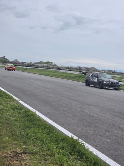 The image depicts a race track in Kaloyanovo, Bulgaria. A black car with "CARSDONE" written on the side is in the foreground, positioned on the track. An orange car is further down the track. The track is bordered by green grass and white lines. The sky is overcast.