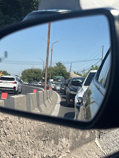 This is a rearview mirror reflection of a traffic jam on a sunny day. The scene is likely in the city of Parakar, Armenia. In the reflection, we see a white SUV in the left lane, with an Armenian license plate. The road is lined with concrete barriers. Behind the SUV, there are multiple lanes of stopped traffic, including cars and a green bus. Overhead power lines and streetlights are visible. The background consists of trees and a clear blue sky. The foreground shows the rough texture of a concrete wall, likely the side of the road or a bridge.