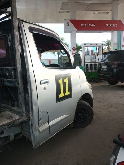 The image captures a street-level view of a Pertamina gas station in Tocina, Indonesia. A silver utility vehicle with the number "11" prominently displayed in yellow on its side is parked at a gas pump. The vehicle appears to be a work truck, with exposed metal framing at the rear. A person, partially visible through the driver's side window, is seated inside the vehicle, facing forward. In the background, a black SUV is parked at another pump. Above the pumps, a sign indicates "BIOSOLAR" and "DEXLITE". The ground is paved with asphalt. The lighting suggests it is daytime.