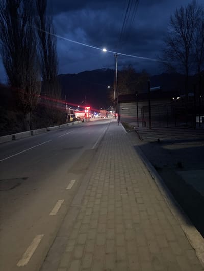 The image displays a roadside scene in Meghri, Armenia, at night or twilight, characterized by an overcast sky with dark, heavy clouds and visible mountains in the distant background. A two-lane asphalt road extends from the foreground into the distance, with a dashed white line marking the right edge of the immediate lane. Parallel to the road on the right, a sidewalk constructed from grey rectangular pavers is present.
A functional streetlight on a utility pole provides bright white illumination in the mid-ground, casting light onto the road and sidewalk. Overhead power lines or communication cables are visible across the upper portion of the frame. On the left side of the road, tall, leafless deciduous trees are present against an unlit, rising landmass. Further down the road on the left, a commercial establishment is illuminated by prominent red and white neon lights. Multiple vehicle headlights and taillights are visible on the road in the distance, indicating traffic, with a large truck discernible near the illuminated commercial area.
On the right, beyond the sidewalk, a low-rise building with a dark roof and lighter-toned walls is visible, fronted by dark, skeletal structures. No individuals are discernible within the frame.