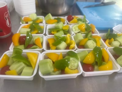 The image shows a close-up, eye-level, outdoor shot of many small, white, rectangular containers filled with chopped vegetables. The vegetables include green bell pepper, yellow bell pepper, cucumber chunks, and cherry tomatoes. The containers are arranged in rows on a metallic surface, likely a table. In the background, a red mug with a white logo, a stack of white disposable bowls, a large metal bowl, and a blue cutting board with a knife are visible. The overall scene suggests food preparation or catering. The location is Namangan City, Uzbekistan.