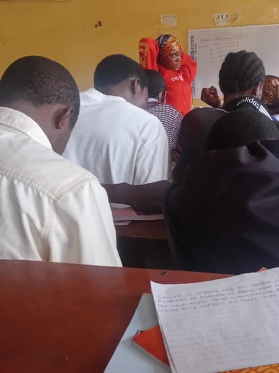 The image depicts an indoor scene, likely a classroom, in Ogbogbo, Nigeria. In the foreground, a wooden desk is visible. On the desk, a sheet of lined paper with handwritten text is placed on top of a stack of books. The text on the paper reads: "Courts in Nigeria rely on various r\nProclamation of Statutes to State rele\nThe law aught to be helm relevant t\nfitles fully explain all there rules".
Several individuals are present in the background, mostly seen from the back or side. One person, wearing a red hoodie and a patterned headscarf, stands near a whiteboard. The whiteboard has handwritten text and drawings, including what appears to be a list of assignments and student registration numbers. Other individuals, predominantly young men, are seated or standing, appearing to be students. Their attire suggests a casual or academic setting. The overall environment appears to be a modest educational institution, with yellow walls and a whiteboard as central features.