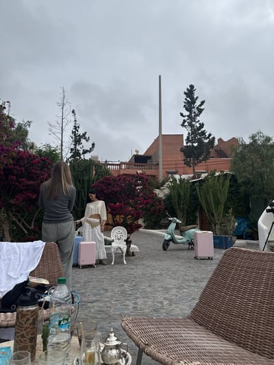 A medium shot shows a scene at Lalla Takarkoust, Morocco, under an overcast sky. A light green scooter and luggage sit on a stone patio. Two women stand amongst flowering bushes. A white metal chair and a tea set are on the patio. Various water bottles and glasses are on a table. In the background are buildings with a unique architectural style and a communications tower.