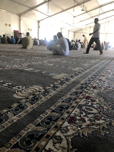 The scene is located in the city of النعيلة, Saudi Arabia, within a large, temporary-looking structure resembling a white event tent with a visible internal metal framework and a high, peaked ceiling. Bright, diffuse natural light illuminates the interior from overhead and the right side, supplemented by ceiling-mounted artificial light fixtures. The floor is entirely covered by patterned prayer carpets in shades of grey, featuring intricate designs in beige, dark blue, black, and accents of maroon.
Numerous adult males, estimated at over thirty, are present. Most are seated on the carpet in rows, facing towards the distant left side of the structure, indicative of a communal gathering or prayer. Their attire varies, including traditional long white thobes and more casual shirts with trousers. In the immediate foreground, a man in a light-colored polo shirt and dark trousers is captured mid-stride, walking from right to left across the carpet. To his left, another man in a white thobe is seated on the carpet, facing away from the camera. Further back, a man in a beige thobe is also seated facing left. The seated individuals appear attentive and still. In the distant left background, two circular fixtures are visible on a plain wall, with a darker, slightly raised area suggestive of a mihrab or minbar. No direct interactions between individuals are clearly observed.
