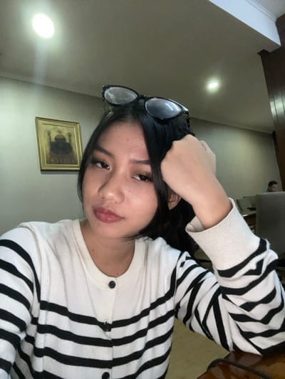 An eye-level indoor photograph displays a young female subject positioned centrally, looking directly at the camera. She has long, dark hair with black-framed eyeglasses resting on top of her head. She wears a white cardigan featuring thick horizontal black stripes and black buttons. Her left hand is raised, with fingers resting against the side of her head, just above her left ear. The background includes a light-colored wall on the left, where a framed picture depicting a dark, domed structure is hanging. Above, two recessed ceiling lights are visible. To the right, the background is blurred, showing additional interior space with indistinct furniture and the partial, blurred figure of another individual seated. A dark, reflective surface, likely a table, is visible in the lower right foreground. The scene is situated in Muka, Indonesia.