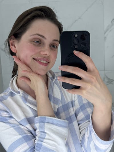 A female individual is depicted in a mirror selfie within an indoor setting in Scafati, Italy, characterized by a white wall with subtle grey marble-like veining. She holds a black-cased smartphone with a visible multi-lens camera array in her right hand, extended towards the reflective surface. Her left hand is positioned with fingers splayed against her lower chin and jawline. She has brown hair, with a braid visible over her left shoulder, and wears a light blue, white, and pink plaid button-up shirt. Her facial expression shows a slight smile, with her mouth slightly open and lips appearing glossy.