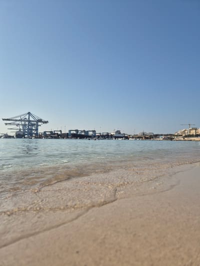 The image shows a wide-angle view of a shoreline with gentle waves lapping onto a sandy beach in the foreground. In the mid-ground, the sea is calm with several boats and a ferry docked. Beyond the water, a busy port area is visible with large shipping cranes, warehouses, and other port infrastructure. The background reveals a cityscape with buildings and more cranes, indicating an urban and industrial environment. The sky is clear and blue, suggesting a sunny day. The overall scene captures a blend of leisure and industrial activity at the port of Birżebbuġa, Malta.
