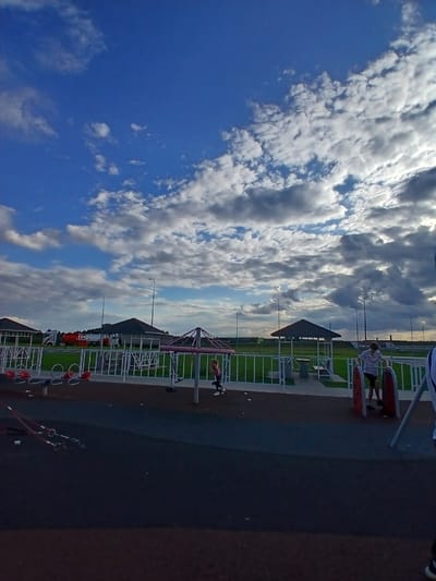 The image depicts an outdoor playground area with several structures and individuals. In the foreground, the ground is paved with dark material, transitioning to a lighter gray in the mid-ground. A white metal fence encloses a grassy area with two hexagonal gazebos, each with a dark roof. To the left of the gazebos, there are red seesaws. Further to the right, a person is interacting with outdoor fitness equipment. In the background, a red and white truck is parked. The sky above is a vibrant blue with scattered white and gray clouds. The scene appears to be located in the city of Vorsha, Russia.