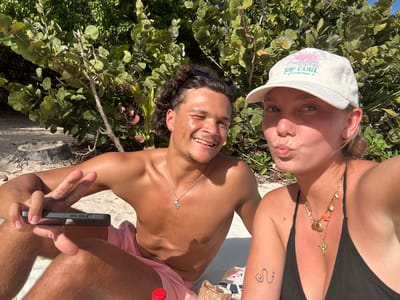 Two individuals, a man and a woman, are photographed in what appears to be a selfie. The man is shirtless with dark curly hair, wearing sunglasses on top of his head and a necklace. He is holding a dark-colored phone in his left hand, making a peace sign. The woman is wearing a white cap with "RIP CURL Guadalupe" printed on it, a black bikini top, and several necklaces. A tattoo is visible on her left arm. They are sitting on a light-colored sandy surface, possibly a beach. Behind them is dense green foliage. A red bottle cap and some debris are on the ground. The location is identified as Sainte-Anne, France.