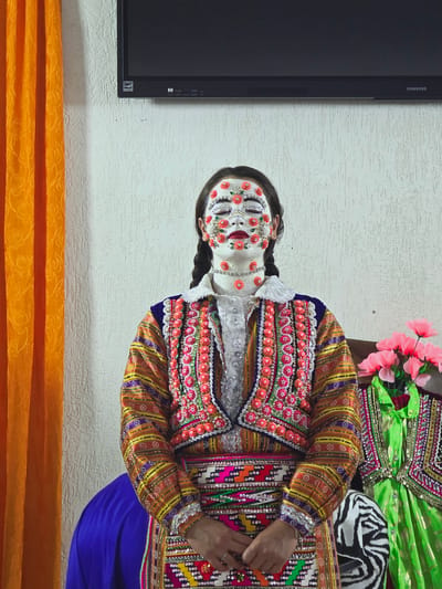 A person with their eyes closed and face painted white with red floral embellishments stands in front of a textured white wall. They wear a brightly colored, ornate traditional garment adorned with numerous small pink and red beads and sequins. The garment features intricate patterns and embroidery. To the left of the person is an orange curtain with a floral pattern. To the right, a bouquet of pink flowers sits next to another green traditional garment. A black television is mounted on the wall above the person. The scene appears to be indoors, likely in Ribnovo, Bulgaria, a village known for its preserved traditional Bulgarian wedding customs.