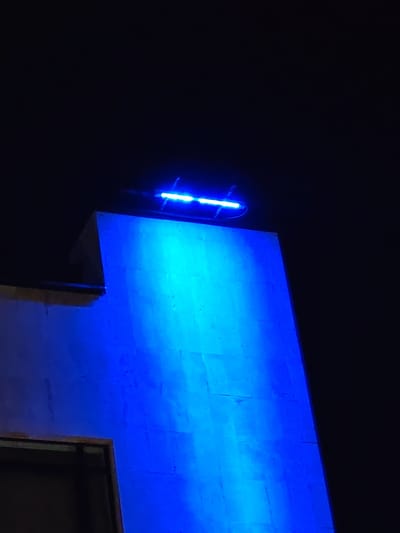 The image captures a nighttime scene in Veliki Preslav, Bulgaria, featuring a large, vertically oriented architectural structure. A linear light fixture, emitting a strong blue light, is mounted horizontally across the top edge of this structure. The blue light casts a vibrant illumination down the facade, highlighting the wall's texture, which appears to be composed of stone or concrete blocks with visible seams. To the upper left, a darker, stepped architectural element is partially visible, receding into shadow. In the lower-left foreground, a dark rectangular opening with a visible brown frame is present. The sky and surrounding areas are uniformly dark, indicating an absence of ambient light beyond the blue illumination. No individuals or other specific objects are discernible.