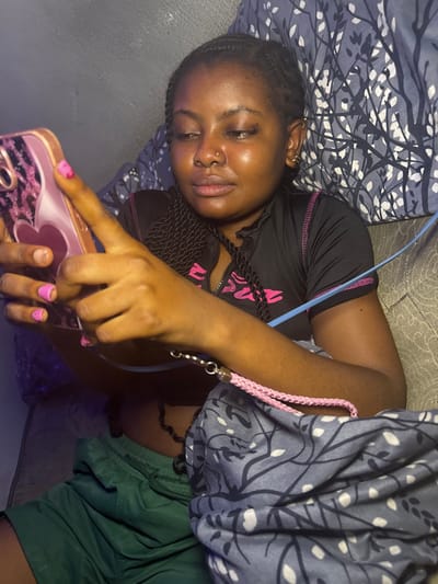 An image taken indoors in Gbongan, Nigeria, depicts a young adult female from the chest up, positioned in a relaxed, reclined manner. She has dark skin, braided hair, a small stud nose piercing on her left nostril, and appears to be wearing false eyelashes. Her fingernails are painted with pink polish, some showing signs of wear. She is dressed in a short-sleeved black top with a zippered front and pink accents, paired with dark green shorts. Both hands hold a smartphone encased in a pink, glittery case with a heart-shaped design. Her right index finger is positioned as if interacting with the phone's screen. A blue cable or cord runs across her left arm and chest, and a pink braided strap with a metallic clasp is visible near her right arm, resting on a patterned blue fabric with white leaf-like designs. This patterned fabric, likely a pillow or blanket, is prominent in the foreground and background, suggesting an indoor setting like a bedroom or living room. The scene is dimly lit by artificial light, with additional illumination emanating from the phone screen.