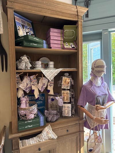 A wooden hutch is filled with various merchandise, including gift boxes, cups, plates, socks, and stuffed animals, all with a lavender theme. In front of the hutch, a mannequin is dressed in a lavender t-shirt and an apron, holding a straw bag. The scene appears to be a retail setting, possibly a shop in Semogorskiy, Russia, judging by the style of the merchandise. The overall impression is one of a charming, rustic store display.