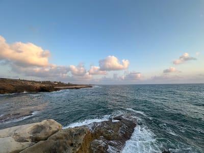 The image depicts a coastline in the Mate Asher Regional Council, Israel. The foreground features rough, textured rocks, some of which are partially submerged by the sea. Waves are actively crashing against these rocks, creating white foam and spray. The sea itself is a deep teal color, with visible ripples and choppiness, indicating a moderate wind.
In the middle ground, the coastline continues with rocky cliffs and a hint of a beach or sandy area further in the distance. Some low-lying buildings and vegetation are visible along the clifftop, suggesting human habitation. The horizon is marked by a clear separation between the sea and the sky.
The sky above is a pale blue, with scattered clouds. Some of these clouds are illuminated by warm, golden light, suggesting it is either early morning or late afternoon, likely during sunrise or sunset. The overall scene conveys a sense of natural beauty and coastal landscape. There are no visible people in the image.