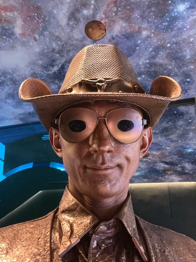 This is a close-up, head-on shot of a bronze statue of a person wearing a wide-brimmed hat with round ear-like protrusions, large circular goggles, and a metallic-textured jacket. The statue has a textured, weathered appearance and appears to be in a seated or tilted position, with its head angled towards the viewer. The background is a dark, starry nebula or galaxy scene, suggesting a space or fantasy theme. The lighting is somewhat dim, highlighting the metallic sheen of the statue. There are no visible text elements. The overall impression is one of a whimsical or fantastical character, possibly from a science fiction or steampunk aesthetic. Given the context of Zakopane, Poland, this statue could be an attraction at a theme park, a sculpture in a public space, or part of a museum exhibit.