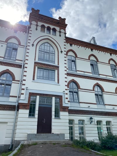 This is a medium-angle, eye-level, full exterior shot of a white building with red brick accents, likely a historical structure in Sortavala, Russia. The building features multiple windows with grey frames, some of which are arched. The architectural style is characterized by brickwork detailing, decorative cornices, and a prominent arched entrance. The sky above is partly cloudy with visible sun glare. There are no people or animals visible in the image. The foreground shows a paved area, a set of concrete stairs leading to the entrance, and some sparse greenery. A downspout runs vertically along the left side of the building.