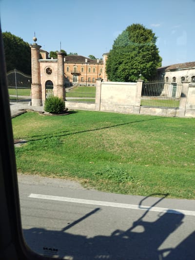 The image depicts a view of a property in Canicossa, Italy, seen from a vehicle. In the foreground, a portion of the vehicle's window frame is visible on the left, and the asphalt road with a white line runs across the bottom of the frame. Shadows of unknown objects, possibly from the vehicle itself, are cast on the road.

Beyond the road, there is a well-maintained green lawn. Further back, a decorative fence separates the lawn from the property. This fence is composed of stone pillars and black metal railings. A segment of a pale yellow wall runs along the fence.

Dominating the background are the architectural elements of the estate. To the left, a grand entrance features two brick pillars topped with spherical finials. Between the pillars is an arched gateway, and above it, a circular window with a decorative grille. A small, dense green bush is planted in front of the gateway. Behind the entrance structure, a large, ochre-colored building with a tiled roof and multiple windows is visible. To the right of this building, a substantial, mature green tree stands prominently. Adjacent to the tree, another building with arched windows and a tiled roof is partially visible, appearing to be part of the same estate. The sky above is a clear, bright blue. The overall impression is of a historical or rural Italian setting on a sunny day.