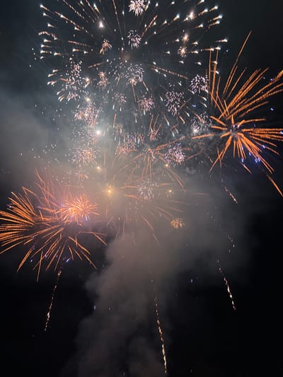 The image captures a nighttime scene of fireworks illuminating a dark sky. Multiple bursts of light, primarily in shades of orange, white, and hints of blue, are visible. Some fireworks appear as scattered sparks, while others form more defined radial patterns. Smoke trails from the explosions drift through the frame, adding to the atmospheric effect. The overall impression is of a celebratory or festive event. The image is a photograph taken during a fireworks display. No specific location in the city of Tutrakan, Bulgaria is identifiable from the visual information in the image.