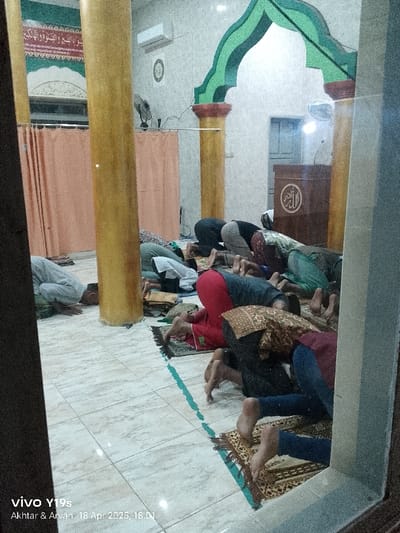 The image shows a scene inside a mosque in Kecamatan Kroya, Indonesia, located at coordinates -6.423 latitude and 107.996 longitude. A group of men are kneeling and bowing their heads in prayer on colorful prayer rugs. They are facing a raised wooden platform with a small white light overhead, a speaker, and a sign with the Arabic word "Allah" displayed. The men are facing a small white sign on the wall behind the platform. There are three large pillars with decorative gold paint visible along with a partial view of a ceiling fan and an air conditioning unit. The walls are light grey and the floor is tiled in a light gray and cream color. A green line separates the tiled area from the prayer rugs. The scene is captured through a glass window with a wooden frame. A phone camera captured the scene and displayed the date and time: "18 Apr 2023, 18.01". The image is slightly blurred due to the window's reflection. A "vivo Y19s" watermark appears in the bottom left corner of the image.