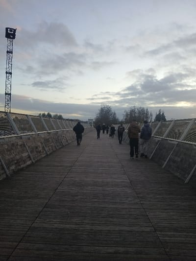 A horizontal shot displays a wooden boardwalk receding into the distance. On either side, metal fences with mesh panels line the pathway. Several figures are walking along the path. The sky is overcast with numerous clouds. A tall metal structure, possibly a crane or tower, stands on the left side of the frame.