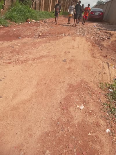 The image depicts a dirt road in Achara Ndiagu, Nigeria. The road surface is red earth and scattered with small rocks and debris. Tire tracks are visible on the road. Five people are standing on the left edge of the road near a parked car. They are of varying heights and are wearing casual clothing. Grass grows along the left edge of the road near some brick structures.