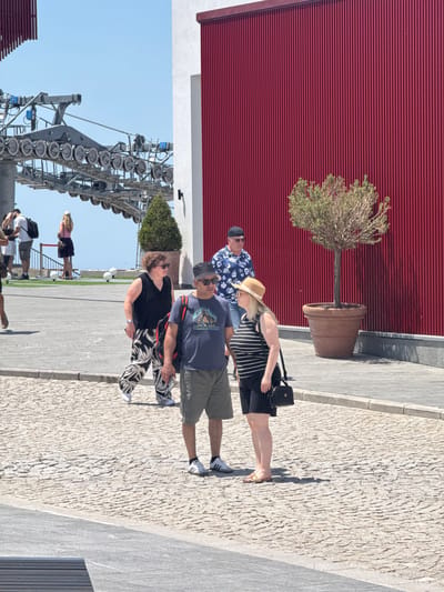 The image depicts a scene in Kuk, Montenegro, featuring a group of people in an outdoor urban setting. In the foreground, a man wearing a dark t-shirt with a graphic and olive green shorts, a black hat, and sunglasses is standing next to a woman in a black and white striped top and black shorts, who is wearing a straw hat and flip-flops. Behind them, a woman with curly brown hair wearing a black sleeveless top and patterned pants walks away from the camera. Further back, a man in a blue floral shirt and a black hat is visible. To the left, part of a cable car structure is present, with its metal frame and numerous circular pulleys. A tall, thin tree in a large terracotta pot stands against a striking red, corrugated metal wall on the right side of the image. The ground is paved with cobblestones. The overall environment suggests a sunny day, possibly a tourist area.