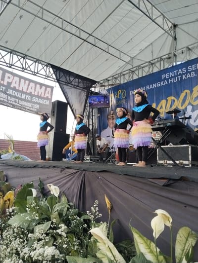 The image captures a scene on a stage, potentially located in Sambirejo, Indonesia. Four young girls stand in a line, each wearing a black top, black leggings, and a multi-layered tutu skirt made of light purple, yellow, and orange tulle. Each has a headpiece or floral crown.

Behind them, on the stage, there are several adults. One man in a white shirt and possibly a formal hat sits on a chair. To the right is a keyboard and a drum set. A television screen hangs above the stage, displaying images. 

Banners surround the stage. One banner reads "PENTAS SENI DALAM RANGKA HUT KAB." Another banner reads "I. PANAMA UT JADULNYA SRAGEN" and includes the name "H. TOMAS HANTIT DARWANTO.SH."

The front of the stage is decorated with white and cream-colored flowers, and green foliage. The stage is draped with a black cloth. The event seems to be an outdoor performance or celebration under a canopy roof.