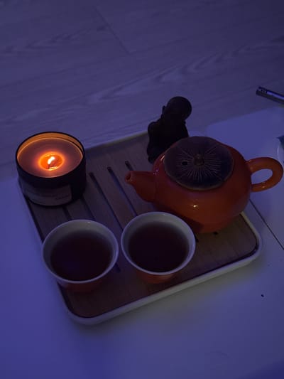 A top-down, rotated view shows an orange teapot and two orange cups filled with dark liquid on a wooden tray with white edging, placed on a light-colored surface. A lit candle in a black container is positioned near the cups. A small black statuette is next to the teapot. The label on the candle is partially visible with Russian text including "KAKAO". The image is taken indoors with mixed blue and yellow lighting. There is no evidence to place the image in Ново-Никольское, Russia.