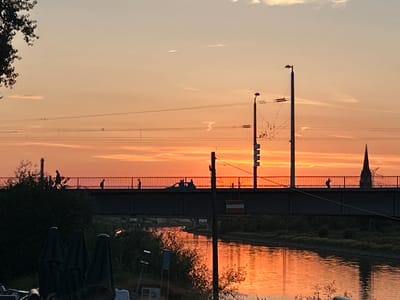 The image captures a sunset over a bridge in Mannheim, Germany, with the Neckar River flowing beneath. Silhouettes of people are visible walking across the bridge, and a car is also present on the bridge. The sky is painted with warm hues of orange and pink, reflecting on the water. Several streetlights stand along the bridge, casting a warm glow. The scene suggests a peaceful evening in the city.