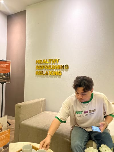 In a hotel lobby in Dumai City, Indonesia, a young person with short, dark hair sits on a beige sofa. They wear a white t-shirt with green trim on the collar and sleeves, featuring the text "I DON'T GET DRUNK MY LIVER DOES" in colorful lettering. The person is looking down at a smartphone held in their right hand, which is resting on their knee. A silver bracelet is visible on their left wrist. To the left of the sofa, a small, light-colored table holds two white teacups and saucers, and a vase of white flowers. Behind the sofa, on a textured wall, golden 3D lettering spells out "HEALTHY REFRESHING RELAXING." To the far left, a brown wall meets a white wall, and a poster on an easel displays the BNI logo and promotional text. The lighting suggests an indoor setting, likely in the evening or a dimly lit area.