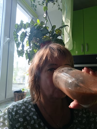 The media file is a close-up, selfie-style portrait of a middle-aged woman in an indoor setting, likely a kitchen. The woman has shoulder-length, reddish-brown hair and is drinking from a clear plastic cup or blender jar filled with a reddish-brown liquid. Her right hand is visible, holding the cup to her lips, with neatly manicured, reddish-pink fingernails. She is wearing a dark green shirt with a white, intricate floral or star-like pattern.
Behind the woman, a large window with white framing and a window handle is visible, offering a view of trees and buildings outside, suggesting it is daytime with bright natural light. Hanging plants, specifically a begonia with large, patterned leaves, are positioned in front of the window and near the woman's head, partially obscuring her. To the right of the woman, there are bright green kitchen cabinets with silver handles. A sheer, pale green curtain is draped between the window and the cabinets.
The overall impression is a casual, everyday moment. The bright natural light and the indoor setting with plants and kitchen elements suggest a home environment, possibly during the day. There is no clear indication of the weather, but the bright light implies it is not overcast. The woman's facial expression is not fully visible due to the cup, but her posture and the act of drinking suggest a relaxed or habitual activity.
Visible text on the cup, though blurry, appears to include "MAX" and possibly a measurement scale. The location context provided is Tugulym, Russia.