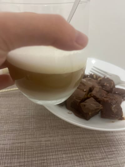 A close-up shot shows a person's hand holding a partially-filled glass cup over a white plate of dessert, possibly brownies. A fork rests on the plate as well. The background includes a textured surface, possibly a table. The location, Mogán, Spain, is not discernible from the image.