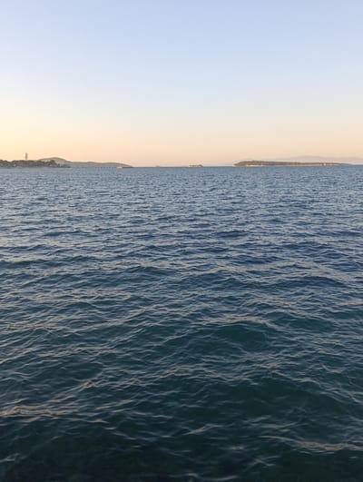 The image depicts a wide expanse of dark blue, rippling water under a clear, pale sky that transitions from light blue at the top to a soft peachy hue near the horizon. In the distance, a city skyline is visible on the left, featuring buildings and a tall tower on a hill. Further along the horizon, several small islands or landmasses covered in greenery dot the seascape, with faint mountain ranges visible behind them on the right. The scene is calm and serene, suggesting a coastal view from Urla, Turkey, possibly during dawn or dusk. There are no people or specific actions depicted in the image; it is a landscape photograph focusing on the sea and distant land.