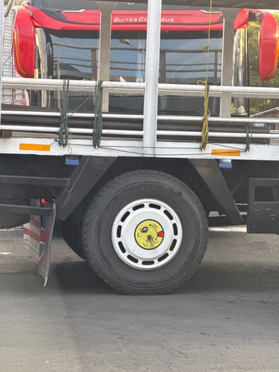 This is a close-up, rotated image of a red and black bus with a smiley face sticker on its tire. The bus is parked on a grey asphalt road. The scene appears to be outdoors during the daytime, with no strong indicators of specific weather conditions.
The subjects are the bus and its tire. The bus has a red exterior with black accents and windows that reflect the surroundings. Text on the bus reads "SUITES C BUS". The tire is black and has a silver rim with a smiley face sticker prominently displayed. The sticker is yellow with a red tongue sticking out and red blush marks, giving it a cheerful expression.
Notable details include the structure of the bus, which appears to be a passenger bus, with visible metal railings and straps. There's a yellow safety marker on the side of the bus. The tire has a visible tread pattern. A portion of what looks like a flag with red and white stripes and blue accents is visible near the bottom right of the image, possibly attached to the bus or nearby.
The overall emotion conveyed by the smiley face sticker is playful and lighthearted, contrasting with the utilitarian nature of the bus. The location context of Melaya, Indonesia, is provided, but not directly visible in the image itself. The image is framed in a way that emphasizes the tire and the smiley face, drawing the viewer's attention to this particular detail on the bus.