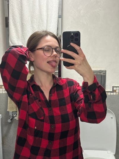 The image depicts an adult female subject taking a mirror selfie inside a bathroom. The specific location within Cava de' Tirreni, Italy, is not discernible from the visual information provided.
The subject is positioned centrally, wearing a red and black plaid flannel shirt. She has light brown hair, appears to be wearing round-framed eyeglasses, and is looking towards the camera (reflected in the mirror). Her right eye is closed in a wink, and her tongue is extended from her mouth. Her left hand is raised and positioned behind her head, while her right hand holds a dark-colored smartphone, featuring a multi-lens camera array and a circular accessory on its back, to capture the image.
The background elements indicate a bathroom environment. To the left of the subject, a chrome heated towel rail with a white towel draped over it is visible, along with a beige towel hanging below. The wall behind the subject features white textured tiles on the upper half, with a decorative border of smaller, square tiles, likely beige or light gray, separating it from a darker tiled lower section. To the right, a white toilet with its lid closed is partially visible. An electrical outlet or wall fixture is present on the tiled wall above the toilet. The lighting is uniform, suggesting an indoor setting.