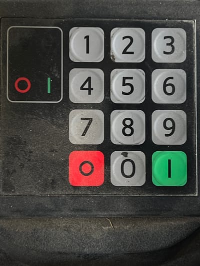 The image is a close-up, overhead view of a weathered, black keypad with numbered buttons and two special function buttons. The keypad is dirty and shows signs of wear. The numbers 1 through 9 are arranged in a standard telephone keypad layout. Below the number 7 is a red button with a circle, likely a cancel or back function. To the right of the red button is a gray button with a zero. To the right of the zero is a green button with a vertical line, likely an enter or confirm function. To the left of the numbered keypad is a blank rectangular area with a red circle and a green vertical line, possibly indicator lights or a small display screen. This keypad is located on a piece of equipment in Lanžhot, Czechia.