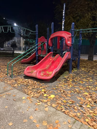 The image depicts a playground in Osorno, Chile at night. The playground equipment includes a red slide with blue railings and green ladder supports. Fallen yellow leaves are scattered on the ground and on the slide itself. The area is dimly lit, with a street light visible in the background. No people are visible in the scene. A concrete sidewalk borders the playground area.