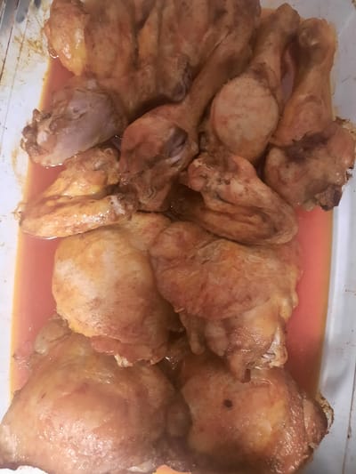 The image shows a close-up, overhead view of a baking dish filled with cooked chicken pieces. The chicken appears to be seasoned and roasted, with some pieces showing browned skin. The dish contains a reddish-orange liquid, likely chicken juices or a sauce, at the bottom. The chicken pieces include drumsticks, thighs, and wings, all arranged somewhat haphazardly in the dish. There is no visible context of a city, including Rosenheim, Germany. The scene is focused solely on the food within the baking dish.