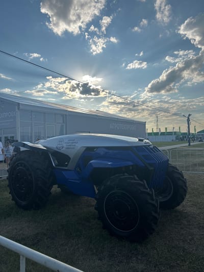 An experimental agricultural tractor, predominantly blue and white with large, rugged tires, is parked on a grassy field in front of a large, modern tent structure. The tent has glass windows and features signage reading "АГРОТЕХПРОЕКТ" (AGROTECHPROJECT) in large, white letters. A smaller sign on the tractor itself indicates it is from "ДОНСКОЙ ГОСУДАРСТВЕННЫЙ ТЕХНИЧЕСКИЙ УНИВЕРСИТЕТ" (Don State Technical University).
In the background, more tents and structures are visible, along with flags and a communication tower. The sky is a clear blue with scattered, fluffy white clouds, and the sun is partially obscured, casting rays through the clouds. A few people are visible in the distance near the tents. The scene suggests an exhibition or demonstration of agricultural technology, possibly at an event in Prutskoy, Russia, given the context of the tractor's origin and the signage. A white railing is in the foreground, partially obscuring the lower portion of the tractor's wheels and the grass.