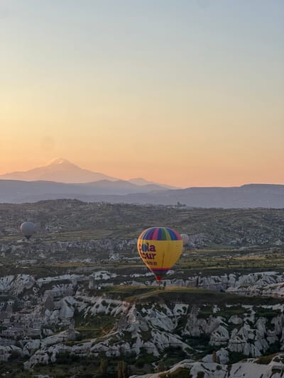 A vibrant hot air balloon, primarily yellow with bands of blue, red, and green, floats prominently in the mid-ground, slightly right of the frame's center. Visible text on the balloon includes "ola hour", "TÜRKİYE", and several Chinese characters. In the lower left background, another, smaller blue-grey hot air balloon is partially visible, also aloft.

The landscape below is a dramatic expanse of unique geological formations, typical of Cappadocia, characterized by light-colored rock structures often referred to as "fairy chimneys." These formations are intermingled with patches of green vegetation, creating a rich, textured terrain. A cluster of buildings, appearing to be a town or village, is nestled among these rocky hills in the mid-right portion of the image.

In the distant background, several layers of mountains or hills rise softly, rendered as hazy, muted silhouettes against the sky. The sky itself displays a beautiful gradient, transitioning from a warm, soft orange and yellow along the horizon, suggesting either sunrise or sunset, to a lighter, ethereal blue-grey higher up. The clear and calm weather conditions are ideal for hot air ballooning, offering a panoramic view of the distinctive landscape. The scene captures a tranquil and majestic moment, likely over Göreme, Turkey, known for its iconic balloon flights.