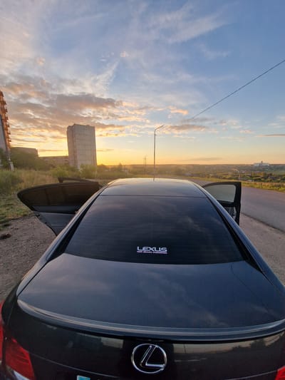 The image shows the rear of a black Lexus sedan parked on the side of a road at dusk in Kashgar City, China. The sky is filled with colorful clouds, illuminated by the setting sun, casting a warm glow over the landscape. In the background, there are buildings and trees, with a prominent tall, concrete building on the left. The car's rear window is tinted, and a "LEXUS" logo is visible on the lower right side of the trunk. The driver's side door is open. The overall scene suggests a peaceful and serene moment during sunset.
